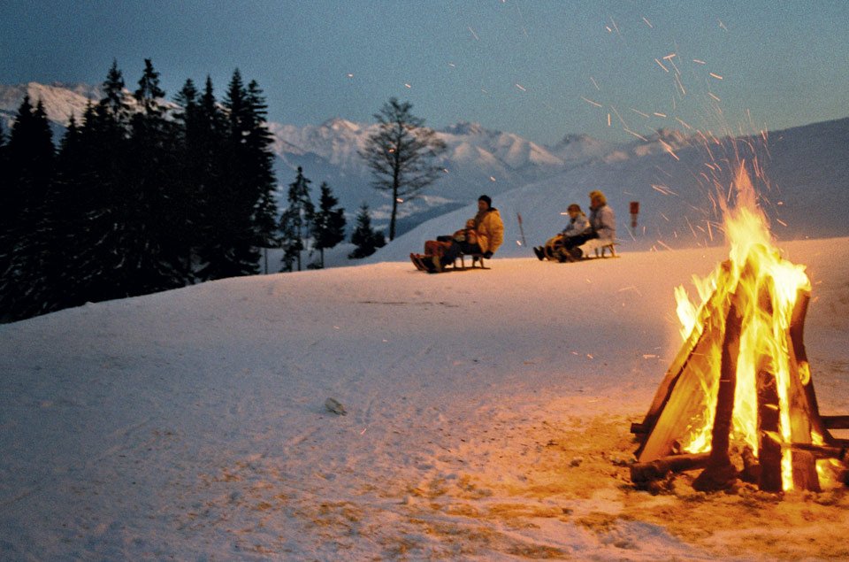 Familie beim Rodeln in der Abenddämmerung, im Vordergrund brennt ein Lagerfeuer